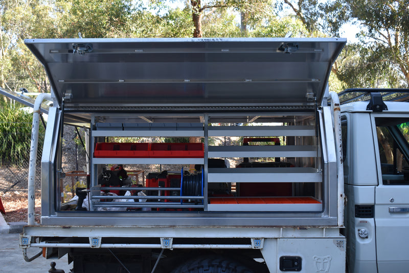 Open truck bed with storage shelves and equipment, surrounded by trees.